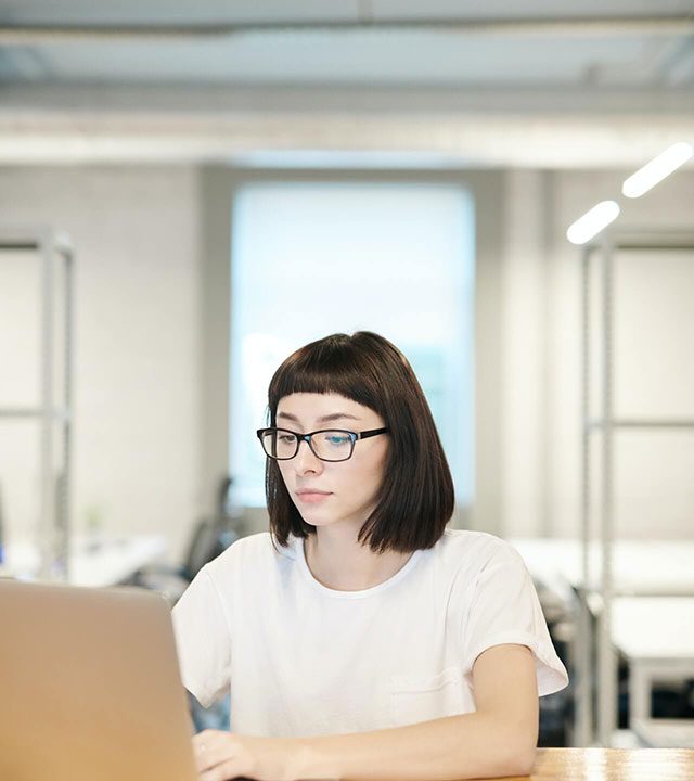 Person wearing glasses and a white shirt working on a laptop at a wooden desk in a modern office setting.