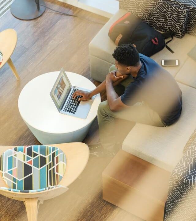Person sitting on a couch using a laptop on a round table in a modern lounge area, with a backpack and smartphone nearby.